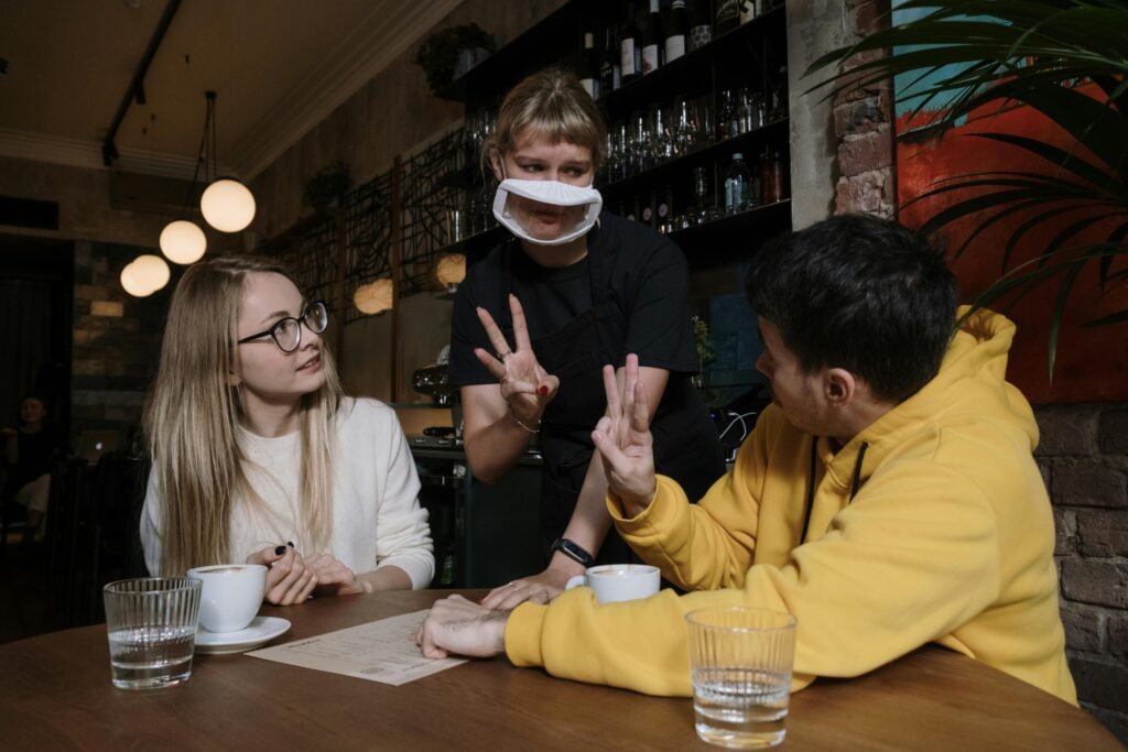 A woman and man use nonverbal gestures with a masked waitress in a cozy café.
